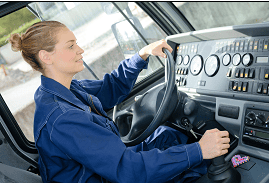 The image captures a young female truck driver focused on the road ahead. She is dressed in a blue work uniform and comfortably seated, hands on the steering wheel and gear stick. The dashboard in front of her features multiple gauges and controls, indicative of a modern truck's cockpit.