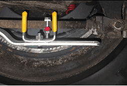 The image displays a brake stroke indicator tool in use, mounted within the wheel area of a vehicle to measure the stroke of the brake chamber. This tool includes a red marker and a caliper-style gauge with yellow handles, set against the backdrop of a brake drum, illustrating the real-world application and importance of precise brake measurement for safety.